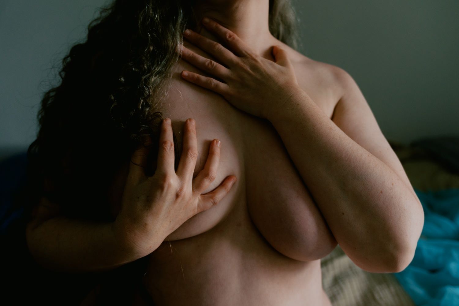Person covering chest with hands in artistic nude photography, soft natural lighting with blue background.