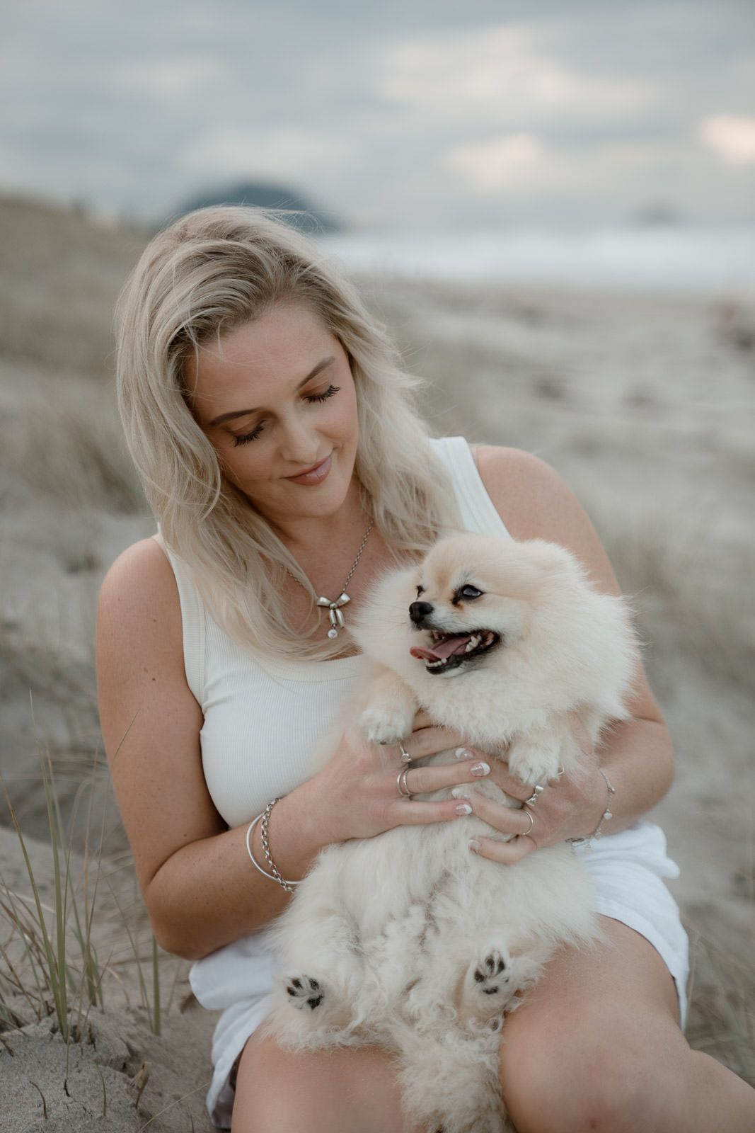 Blonde woman in white dress holding a fluffy cream Pomeranian dog on a sandy beach at dusk.