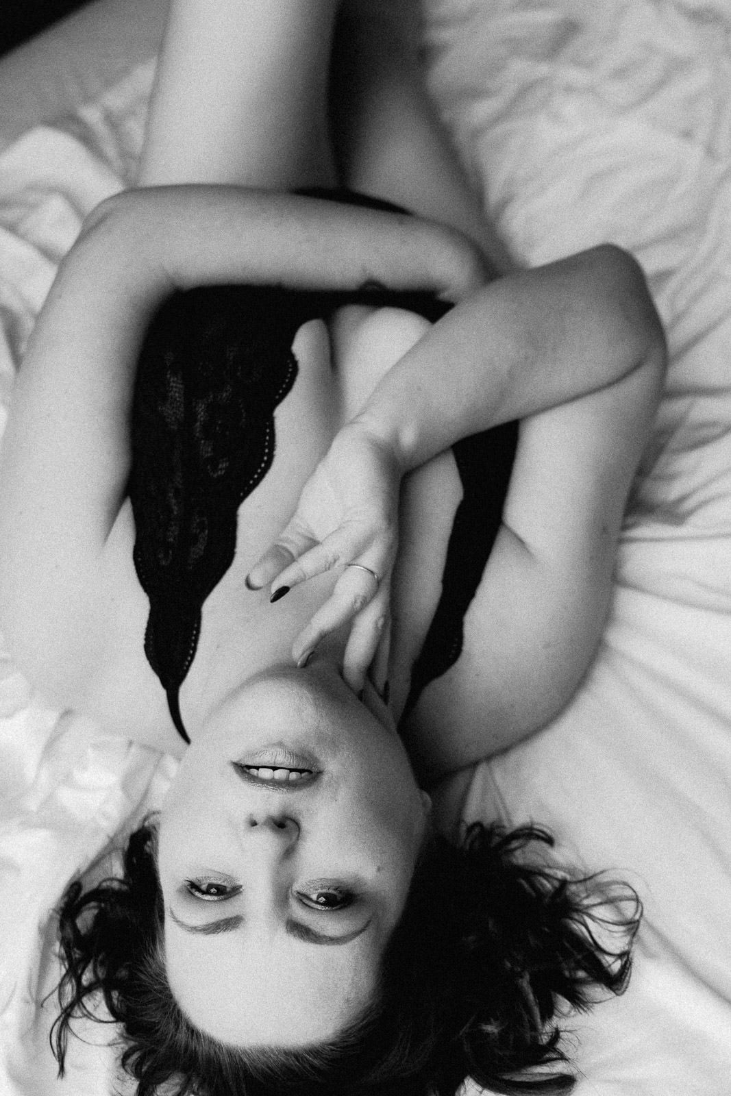 Black and white boudoir photo of a woman in black lace lingerie lying on white bedding.