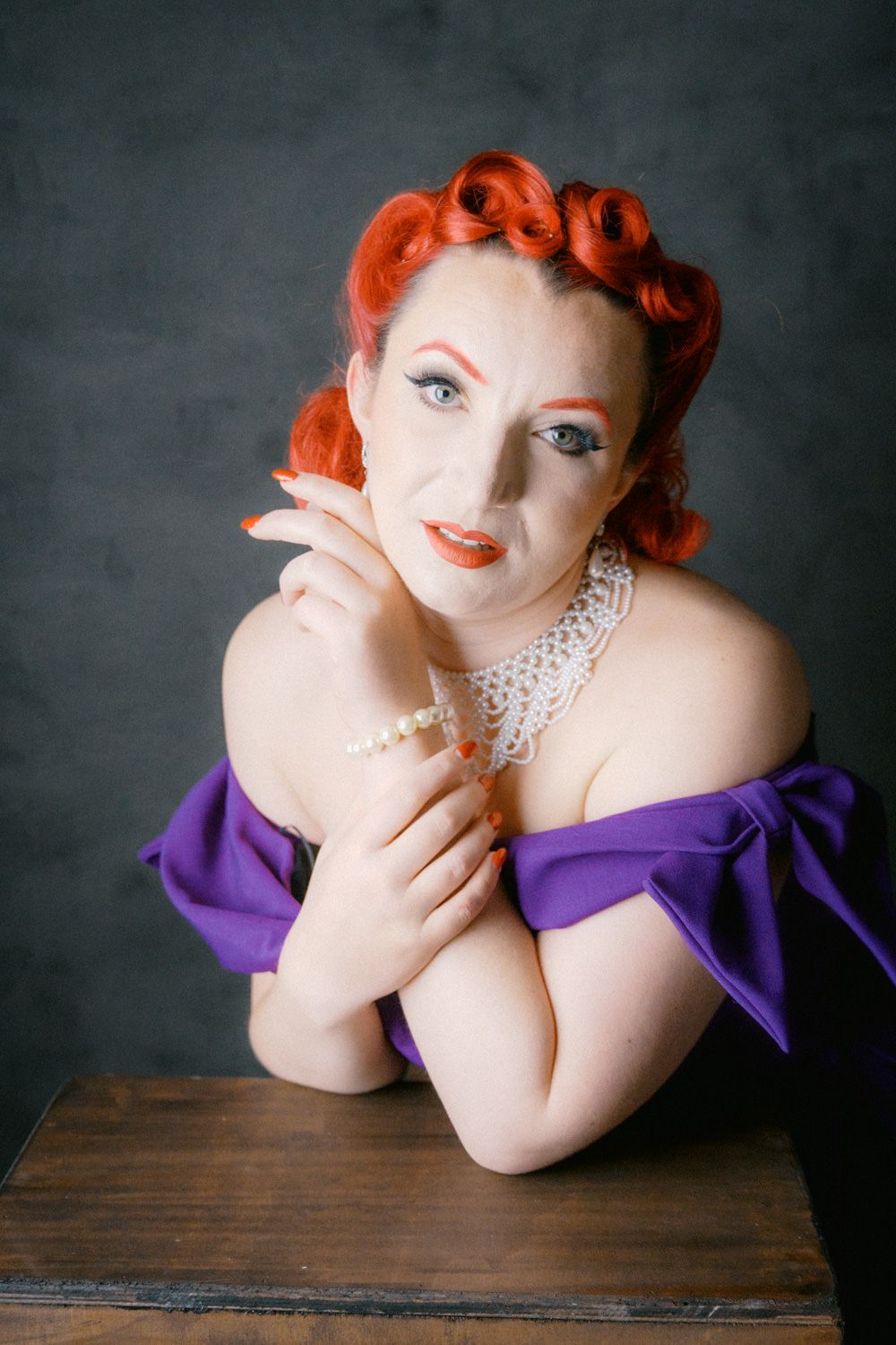 A woman with red pin-up hair, pearl jewelry, and a purple off-shoulder dress poses elegantly at a wooden table.