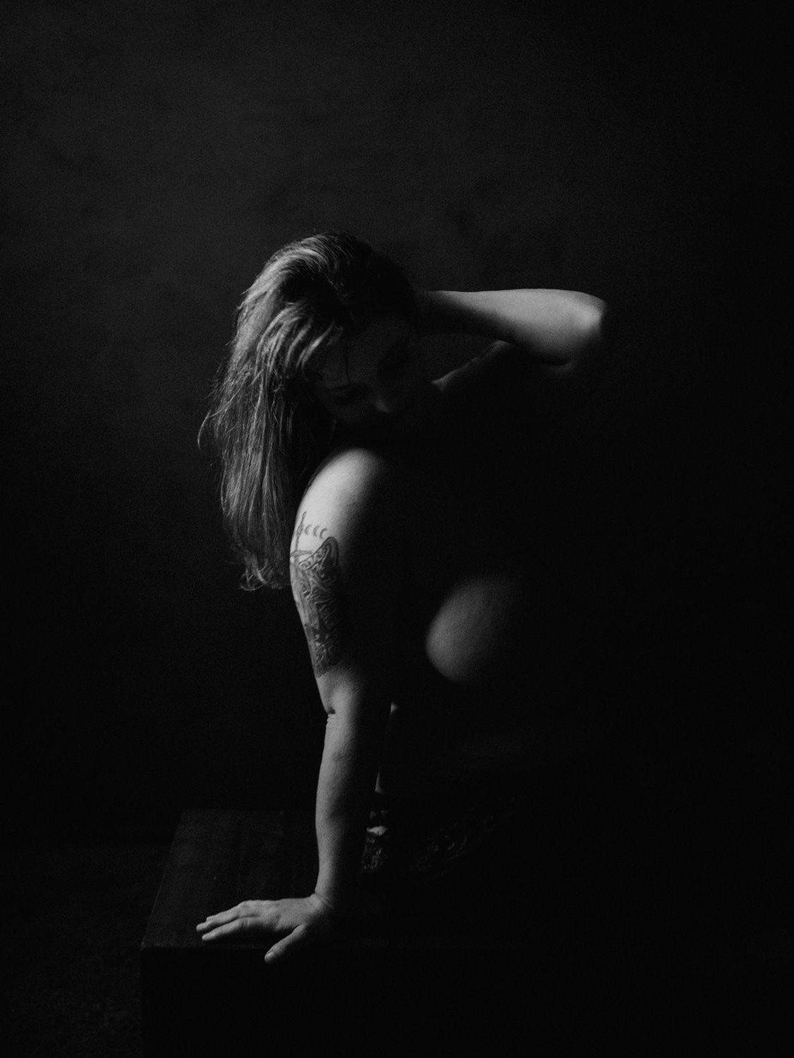 Black and white artistic photo of a woman with long hair and arm tattoo posing in dramatic low-key lighting.