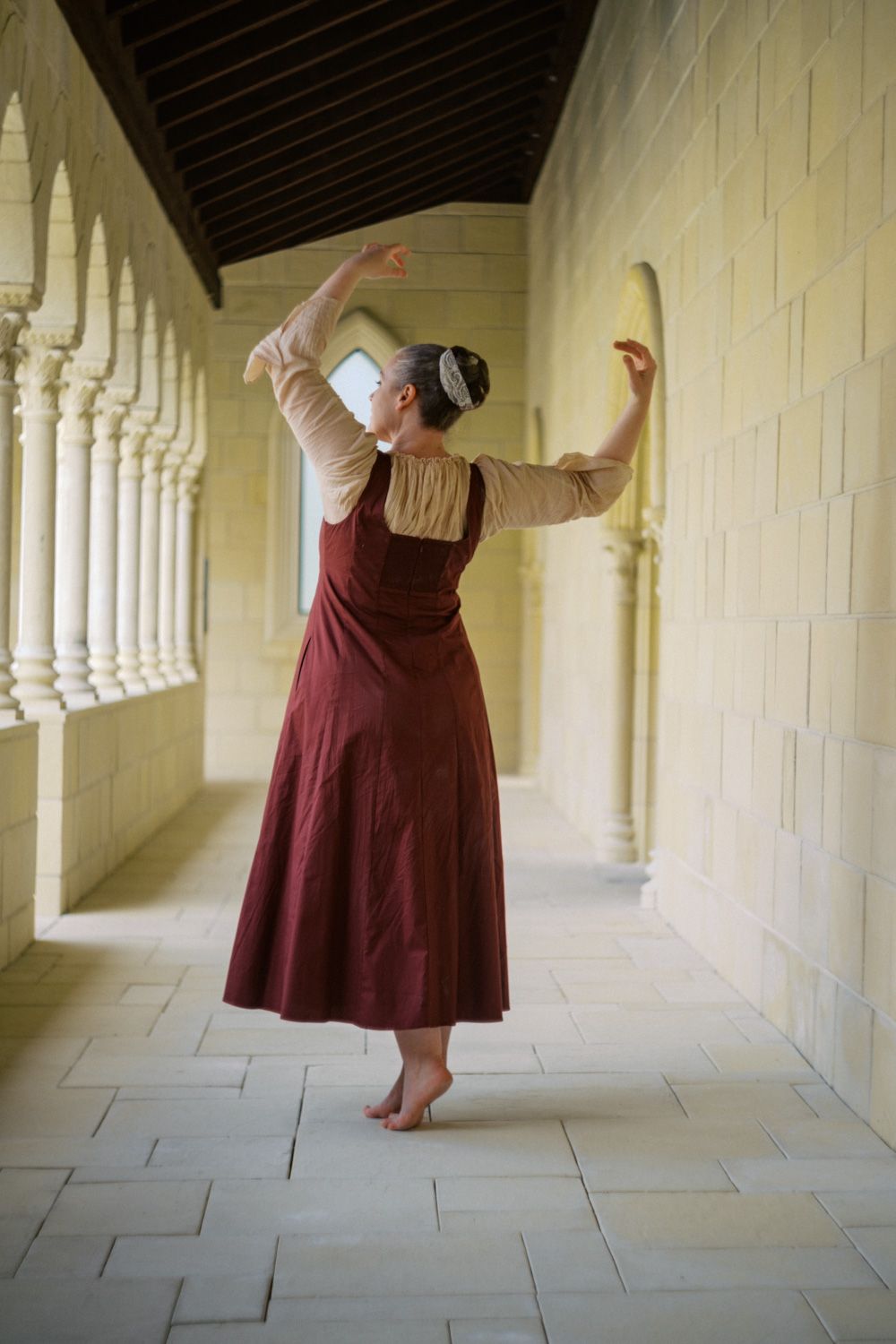 A barefoot dancer in a red dress poses gracefully in a stone archway corridor with white columns.