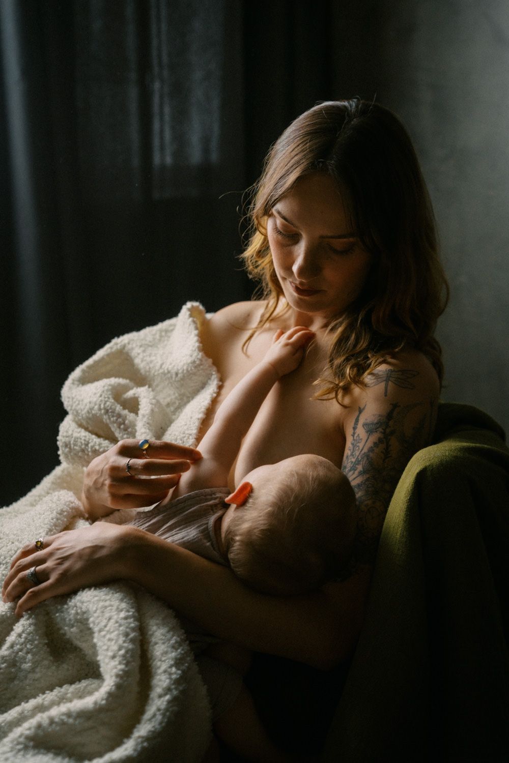 A tattooed mother tenderly breastfeeding her newborn baby while seated in a chair by a window.