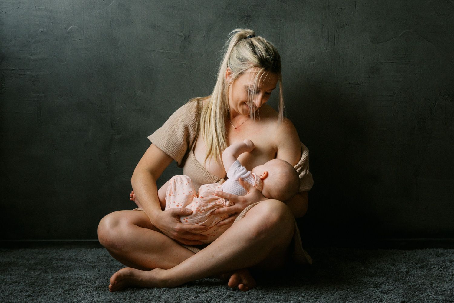 Mother breastfeeding newborn baby while sitting cross-legged on floor against dark gray wall.