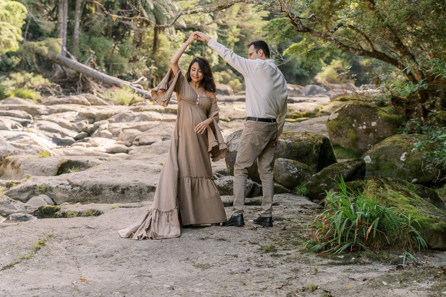 Couple dancing outdoors on rocky terrain surrounded by lush trees, woman in flowing tan dress, man in casual gray outfit.