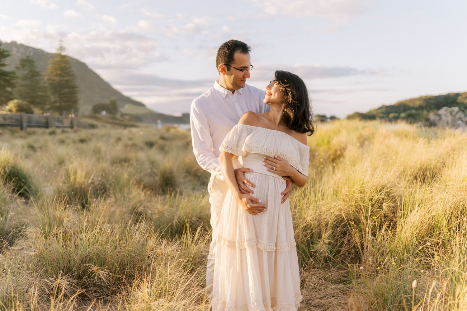 Couple embracing in sunlit field, woman in white maternity dress at golden hour.