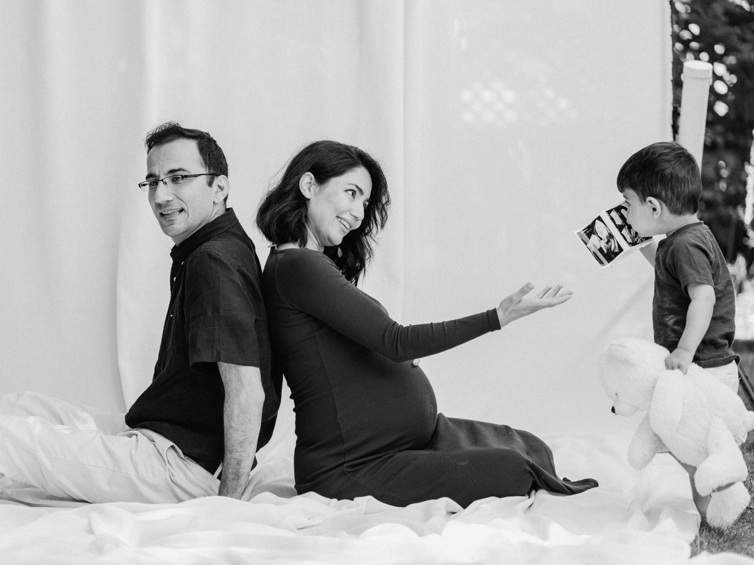 Black and white maternity photo of pregnant couple sitting back-to-back while toddler shows ultrasound photo.