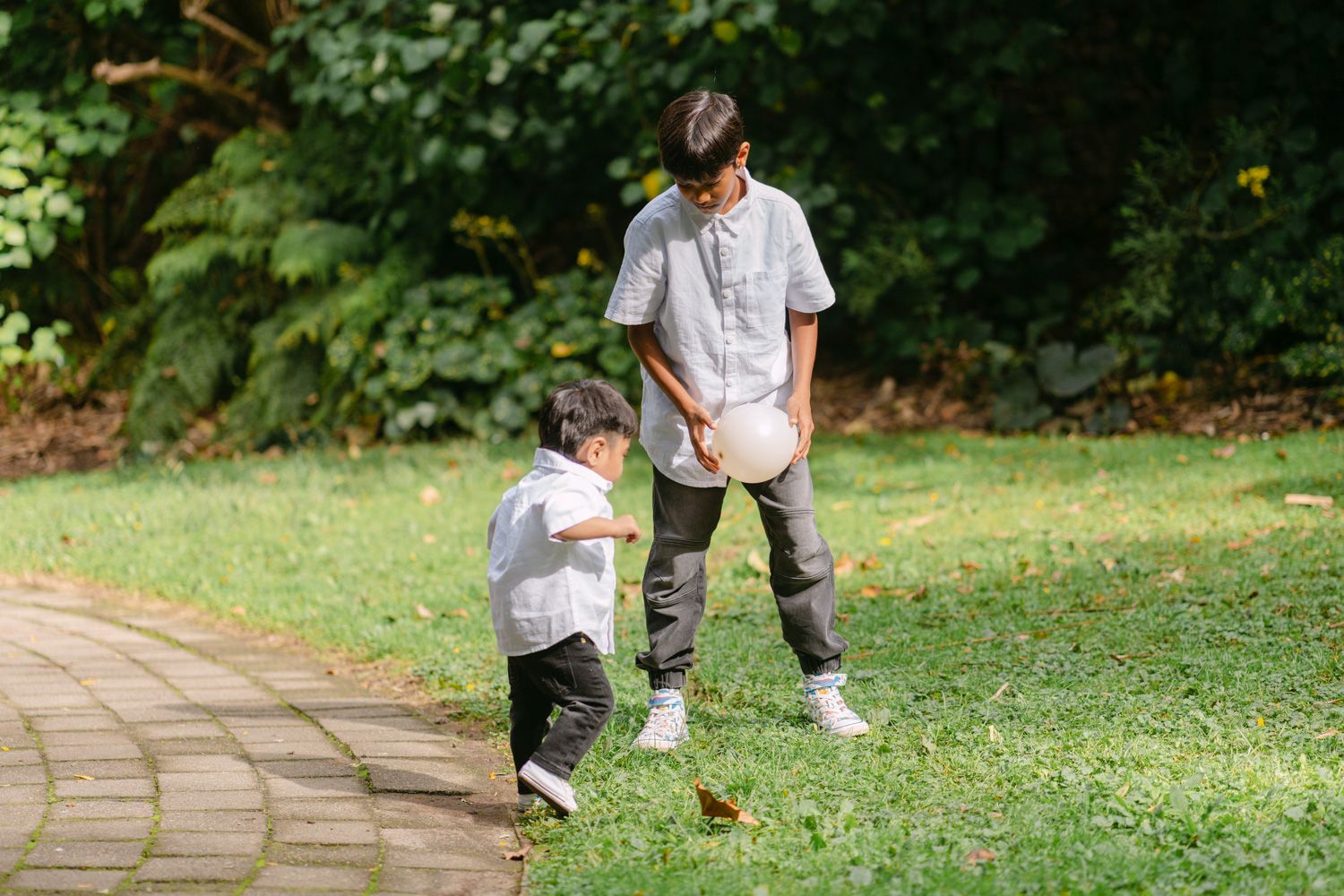 Two boys playing together on a grassy park path surrounded by lush green trees on a sunny day.