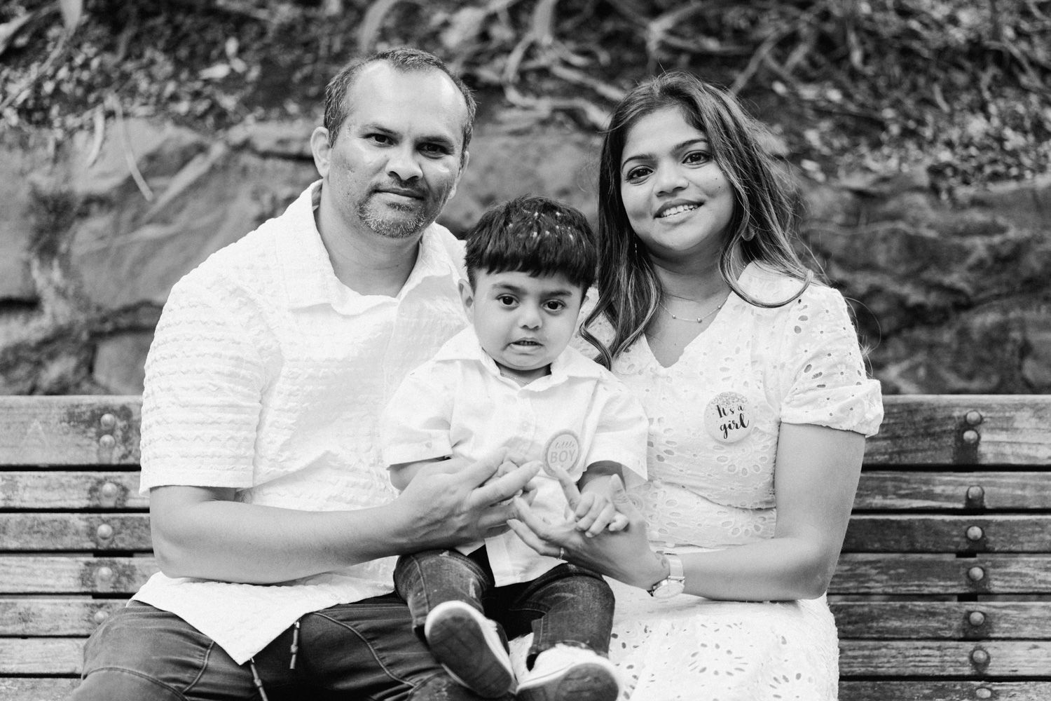 Black and white family portrait of parents with toddler son sitting on bench outdoors.