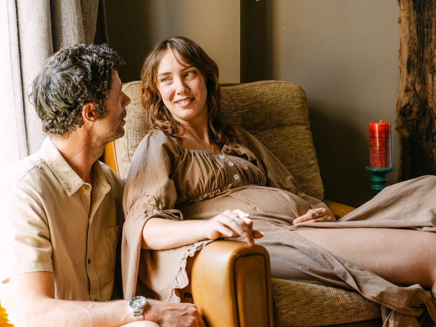 A couple shares a warm moment, woman relaxing in a yellow armchair while man sits beside her smiling.