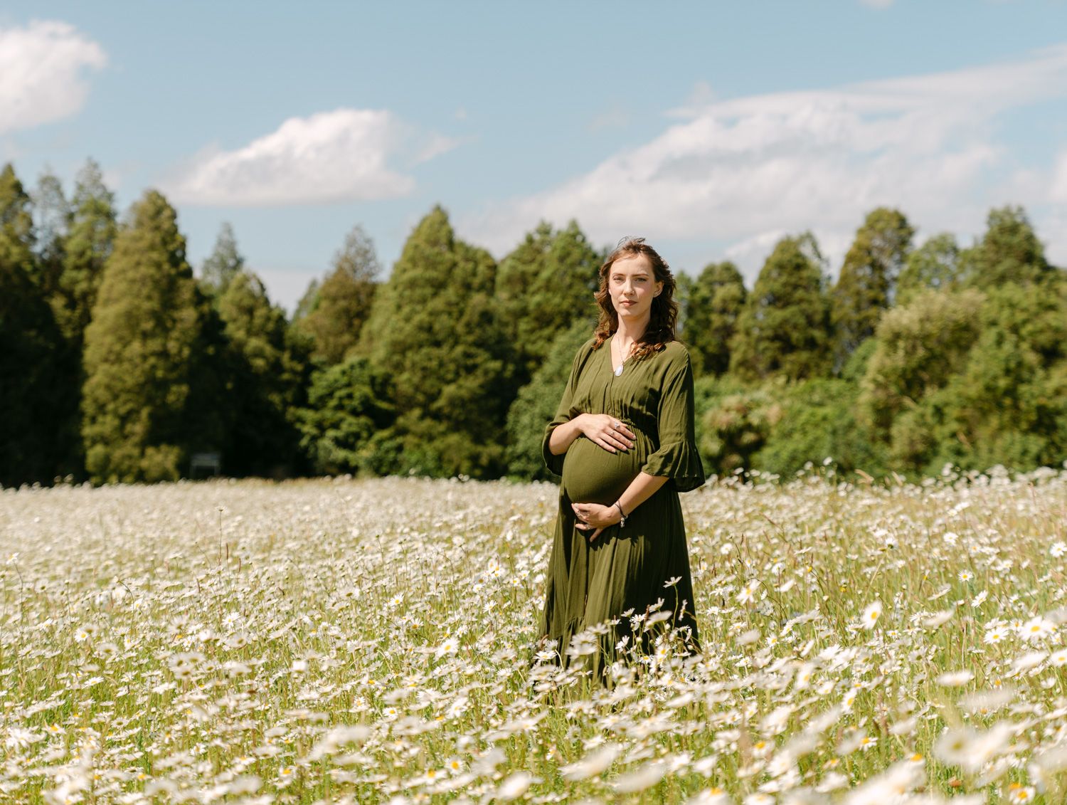 Pregnant woman in green dress standing in a daisy field with trees in background on a sunny day.