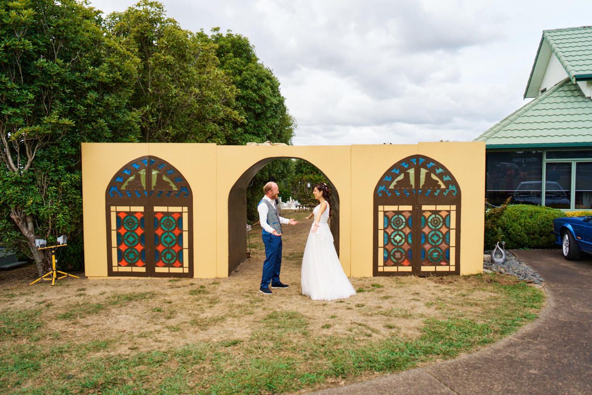 Bride and groom share a first look moment in front of a yellow wall with colorful stained glass arch windows outdoors.