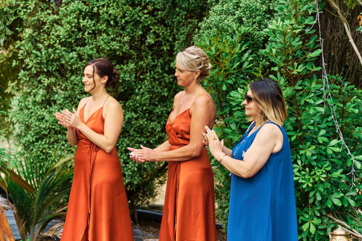 Two bridesmaids in rust orange satin dresses clap alongside a woman in blue at an outdoor wedding ceremony.