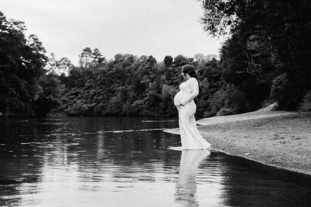 Pregnant woman in white dress standing at river's edge in black and white maternity photo surrounded by lush trees.