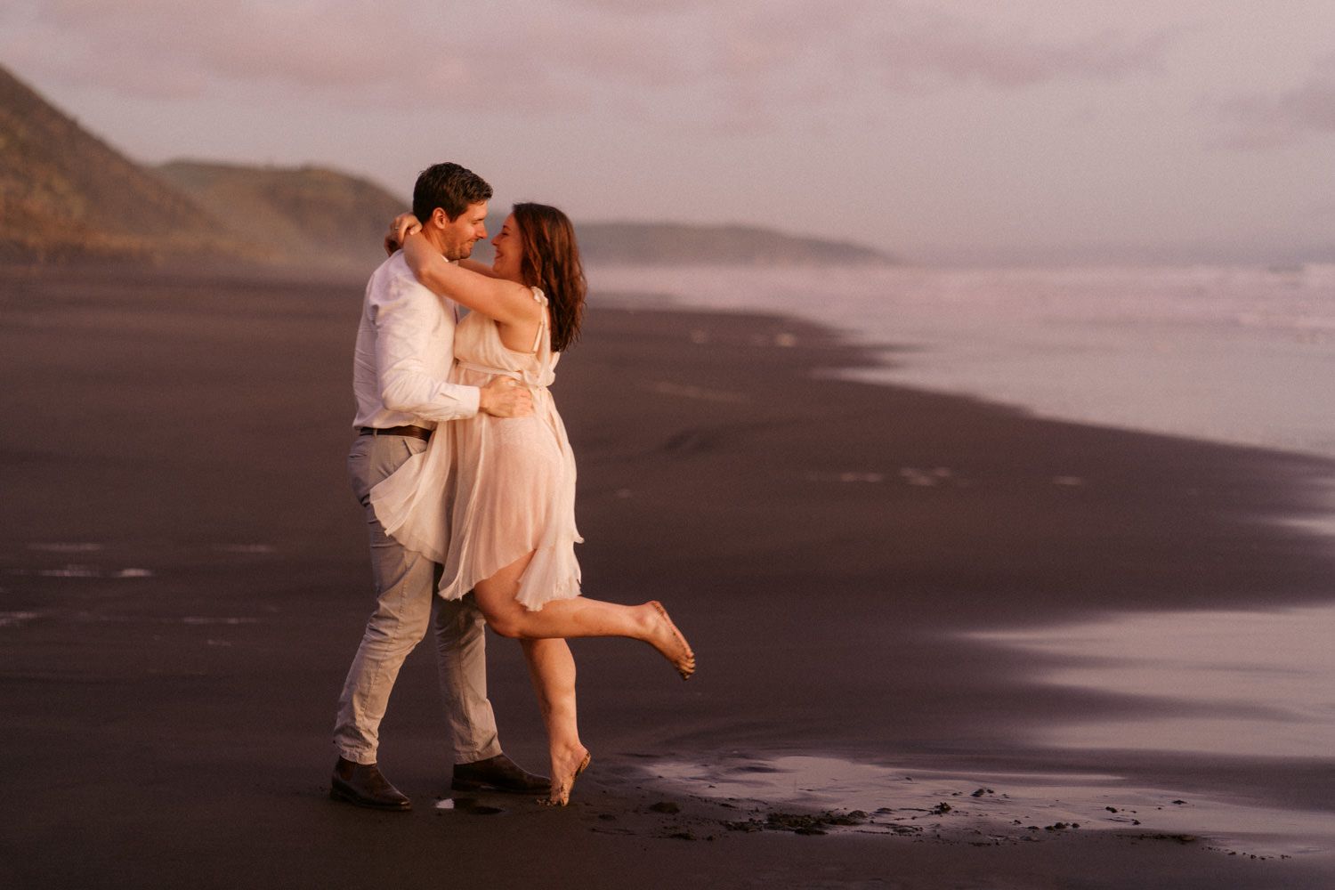 Romantic couple embracing and kissing on a dark sandy beach at sunset with ocean waves in the background.