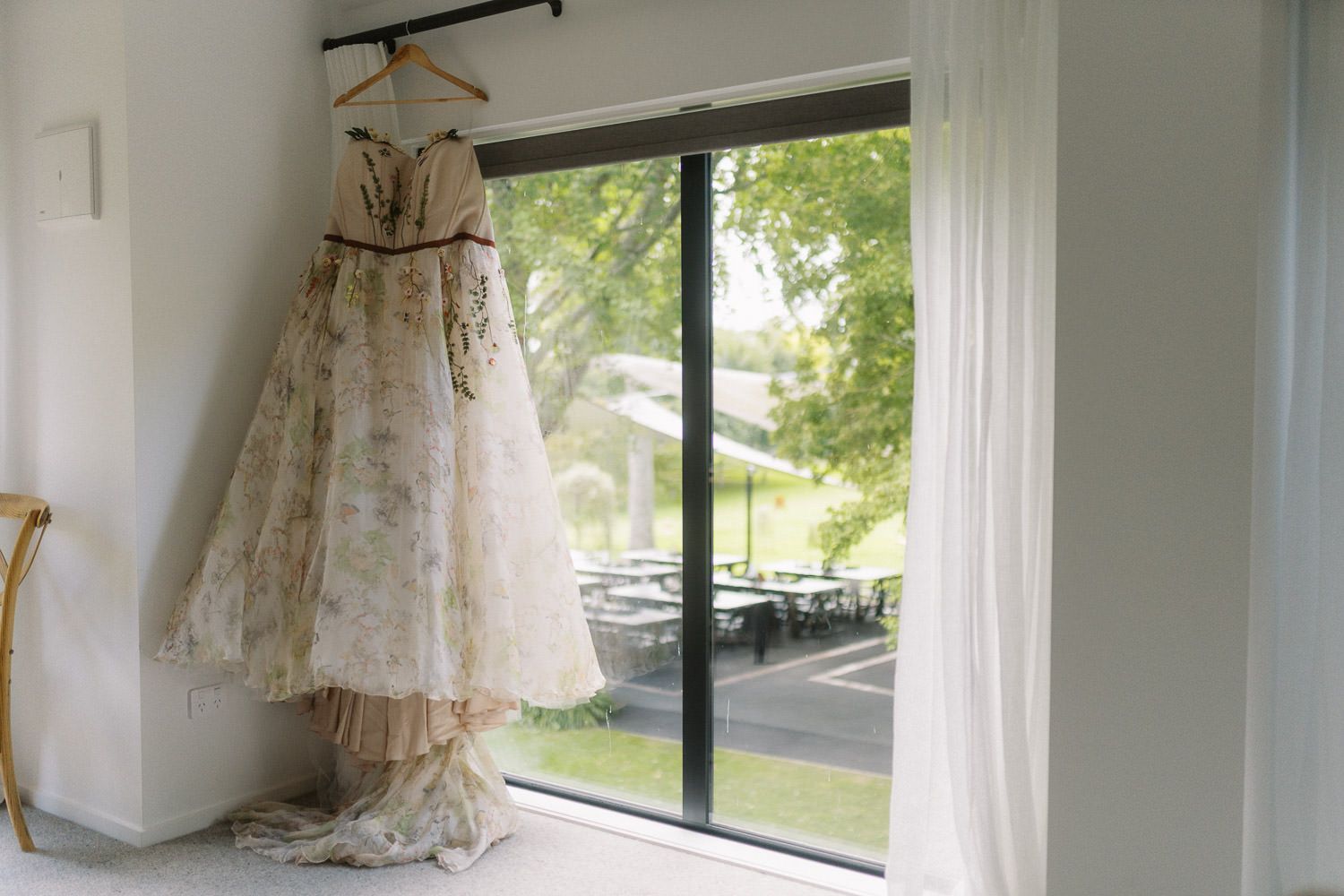 Elegant floral wedding dress hanging by a large window with natural light and garden view.