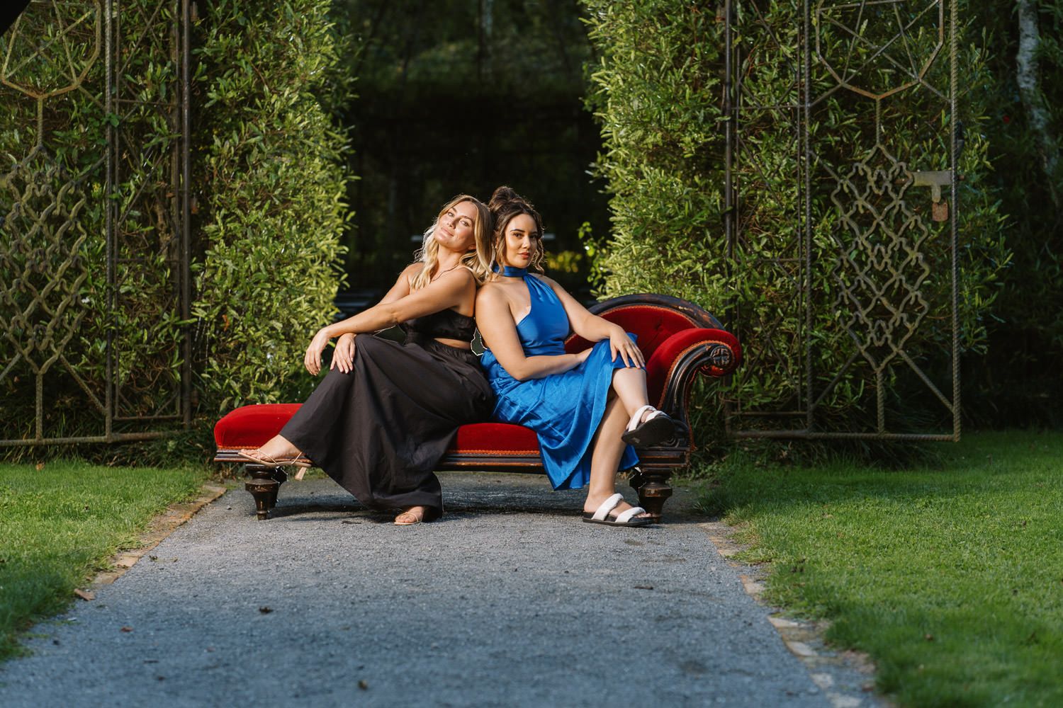 Two women in elegant gowns pose on a red velvet sofa outdoors surrounded by lush green hedges.