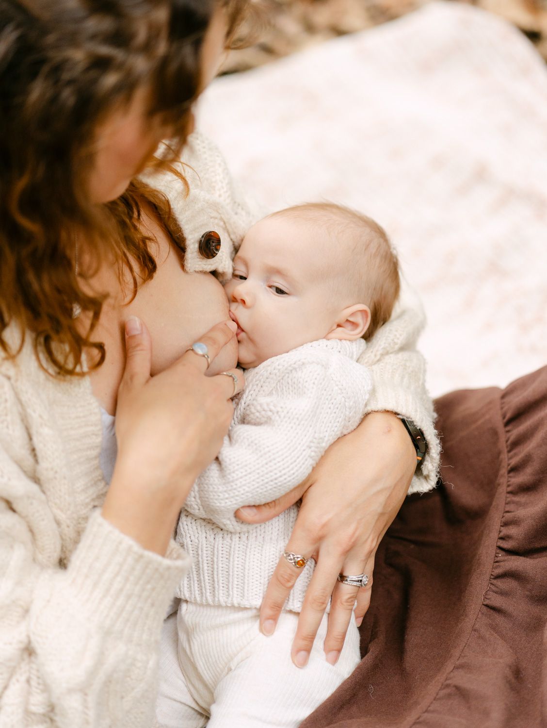 A mother in a cream sweater tenderly breastfeeds her newborn baby dressed in a white knit outfit.