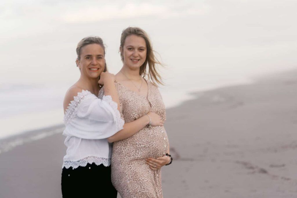 Two women pose on a beach, one in a white top and one in a floral maternity dress, smiling together.