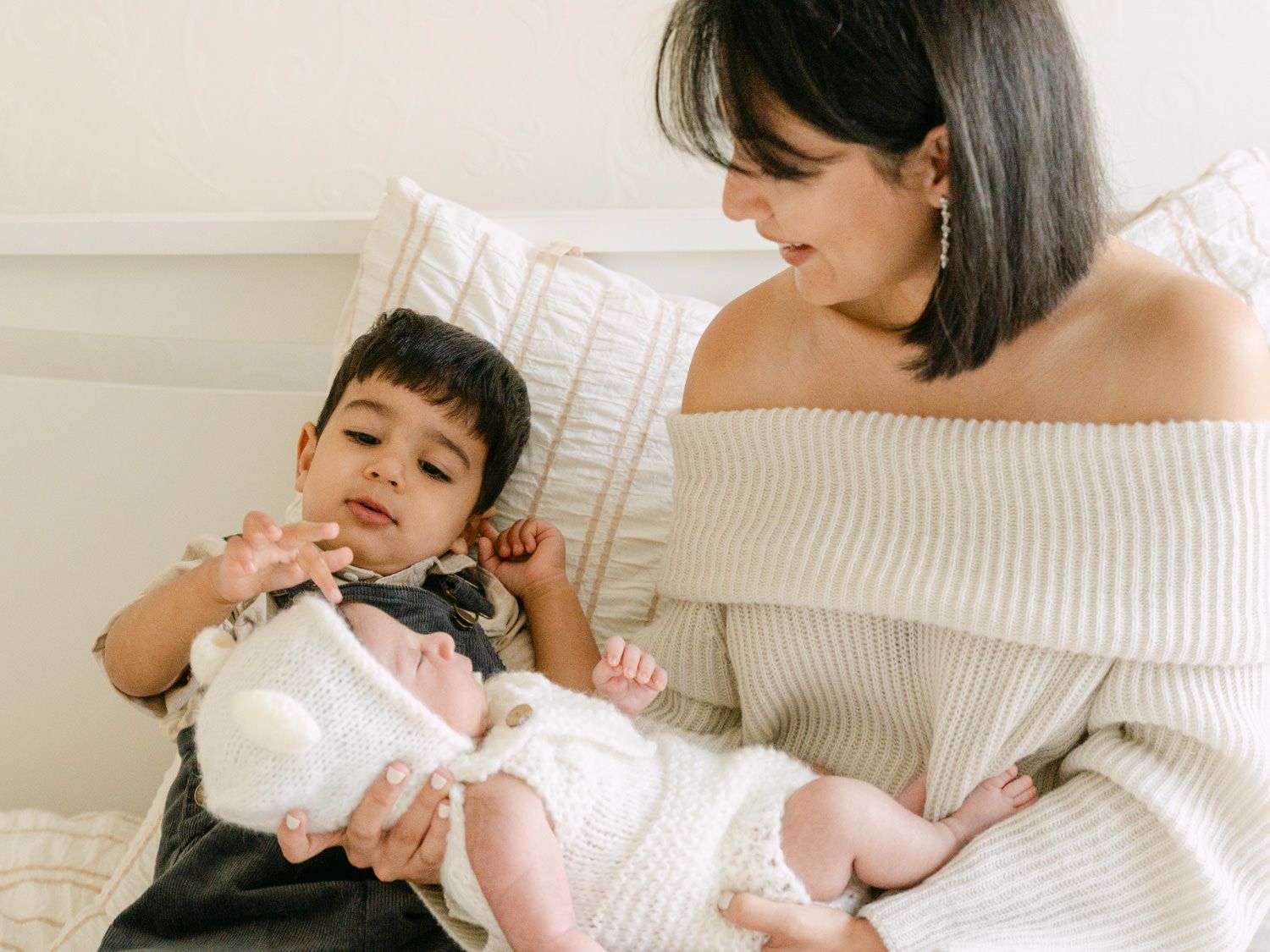 Mother in off-shoulder sweater sits with toddler son gently touching newborn baby sibling.