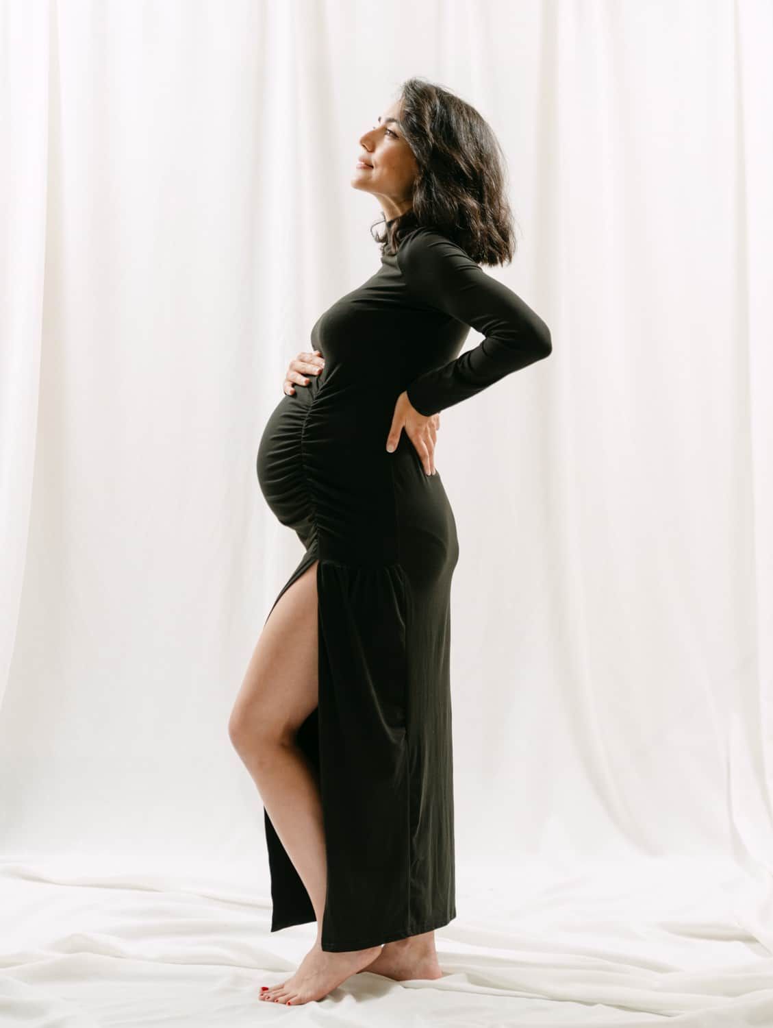 Pregnant woman in black ruched long-sleeve maxi dress with side slit, posing sideways against white curtain backdrop.