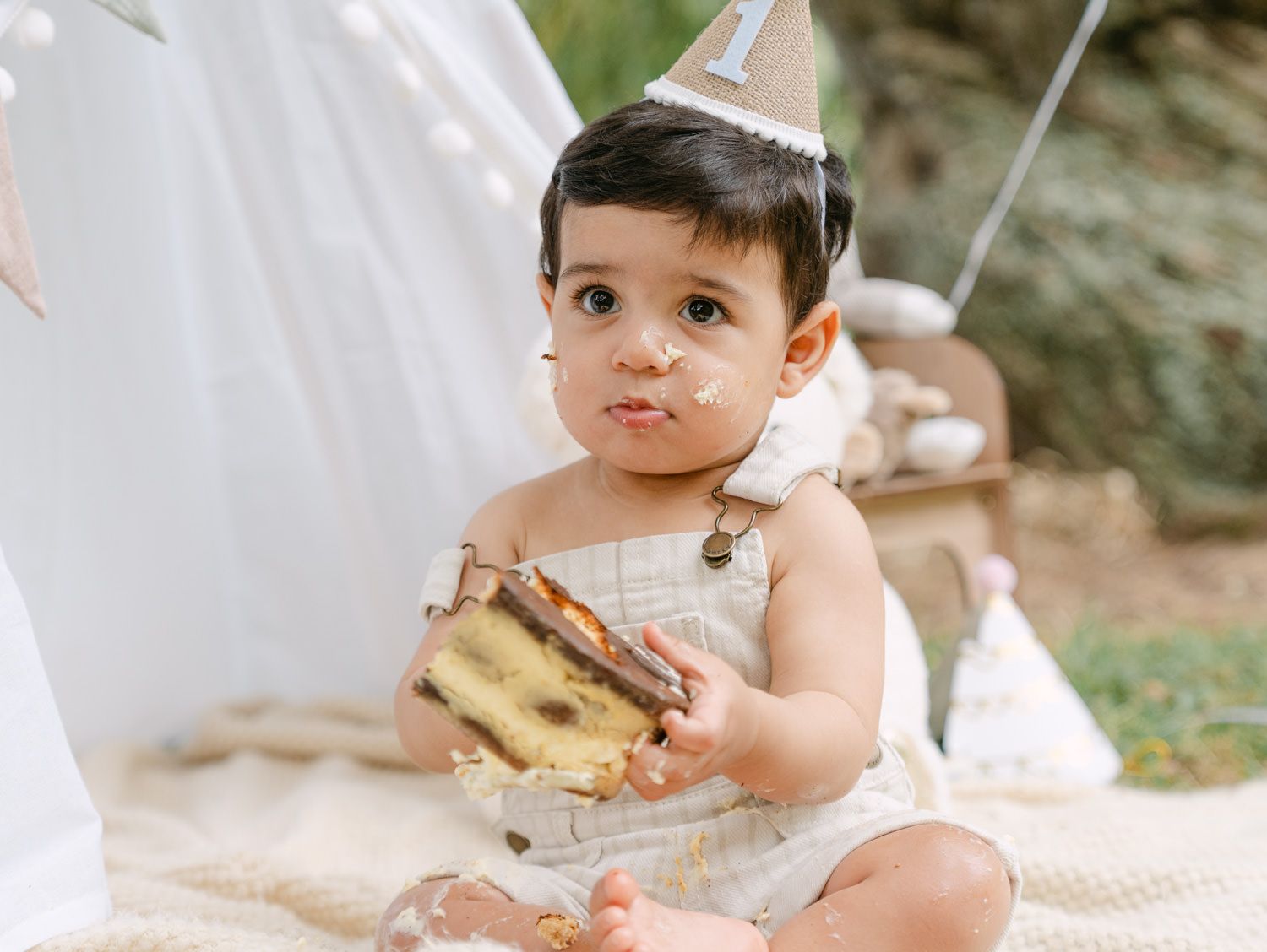 Baby in a party hat enjoys smashing their first birthday cake outdoors during a boho-themed cake smash photoshoot.
