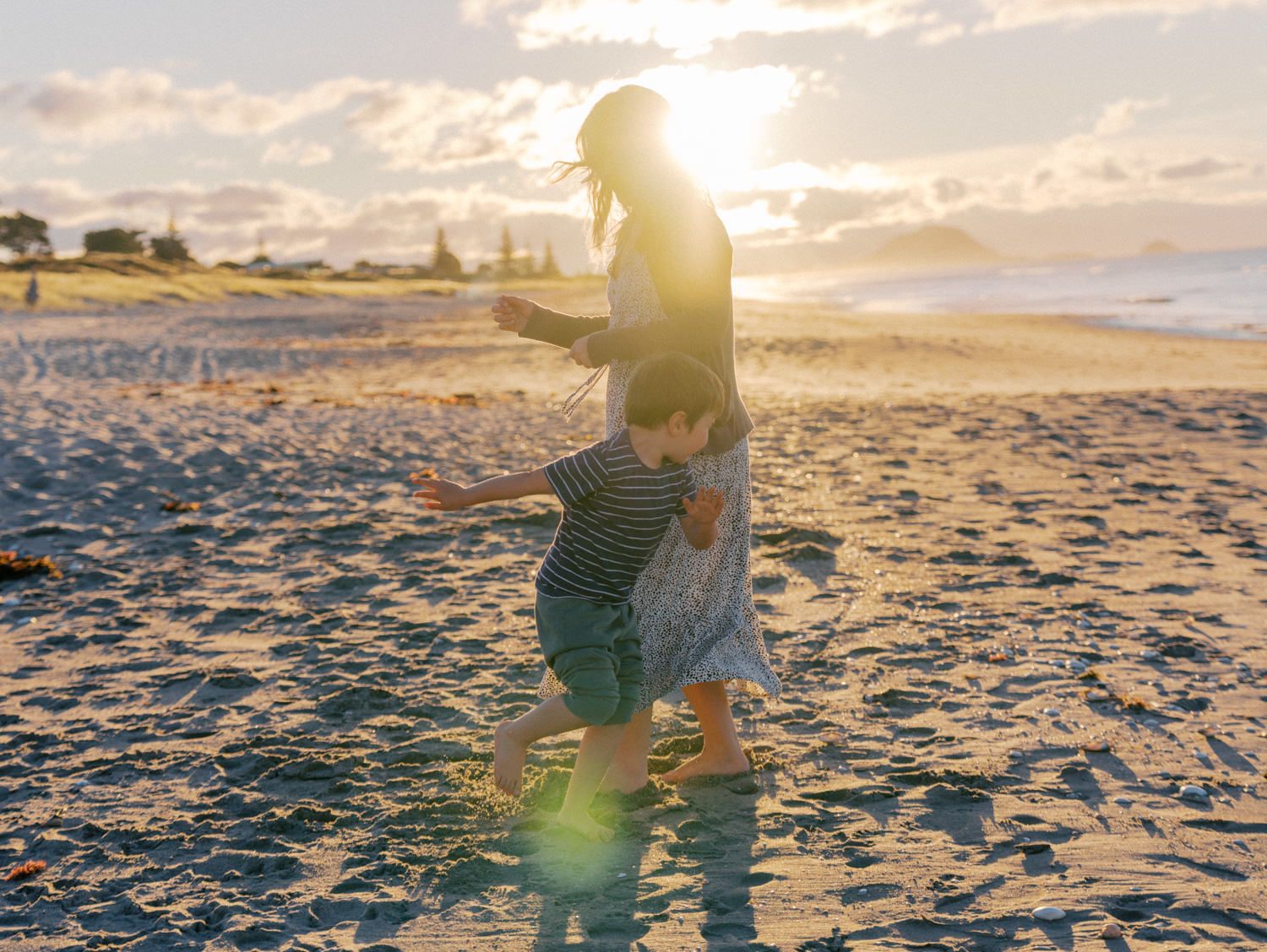 Mother and child playing on a sandy beach at golden sunset with ocean waves and warm sunlight glowing behind them.