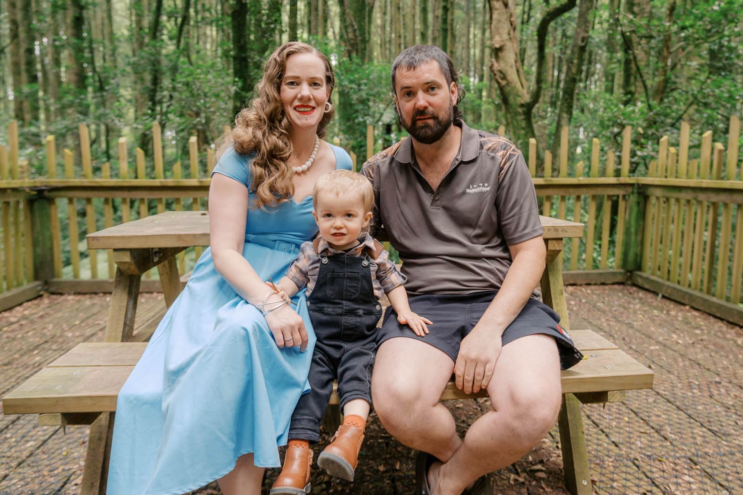 Family of three sitting on wooden bench in forest, parents smiling with toddler in brown boots between them.