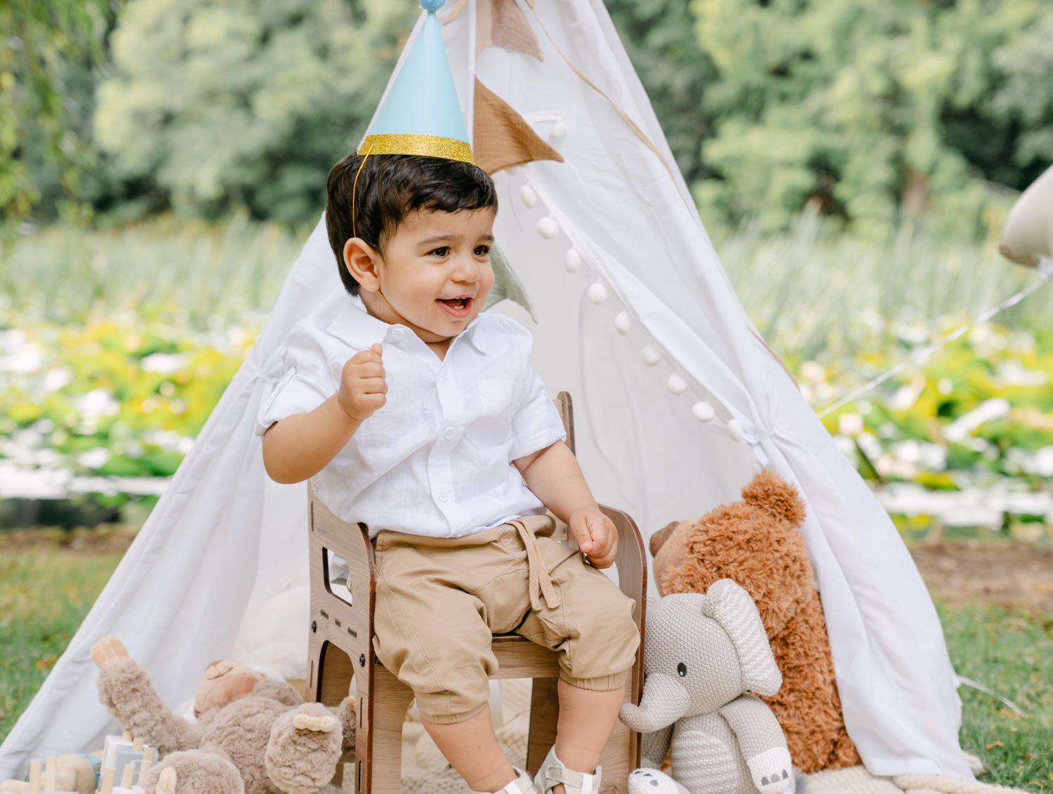 Toddler boy in white shirt and party hat sits by a teepee tent surrounded by stuffed animals outdoors.