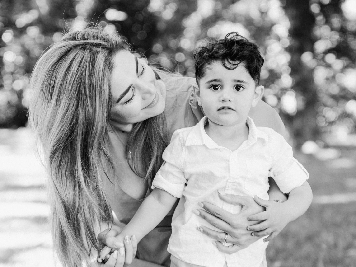 Black and white photo of a smiling woman hugging a young toddler boy outdoors in a park setting.