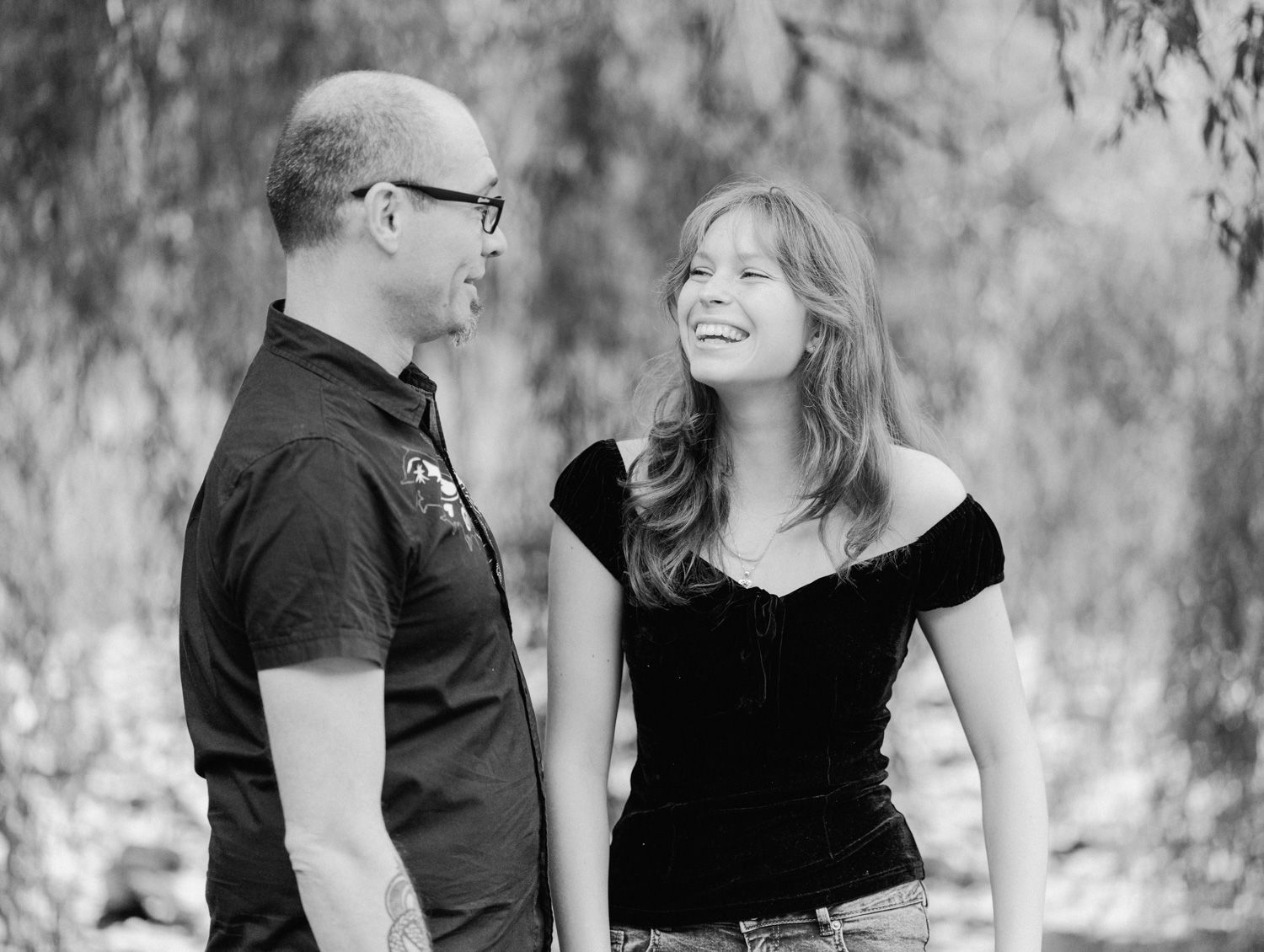 A man and woman sharing a laugh together outdoors in a black and white portrait photo.