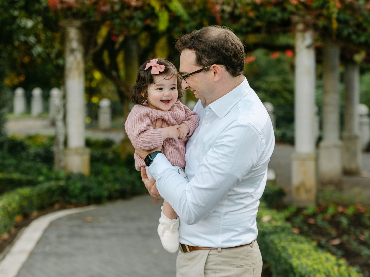 Father holding baby girl in pink outfit at garden with stone columns and lush greenery.