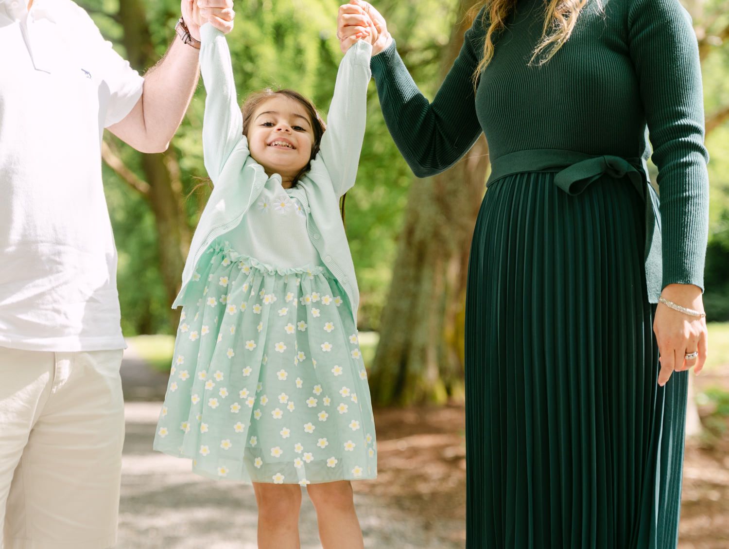 A joyful young girl in a mint polka dot dress holds hands with her parents outdoors in a bright green setting.
