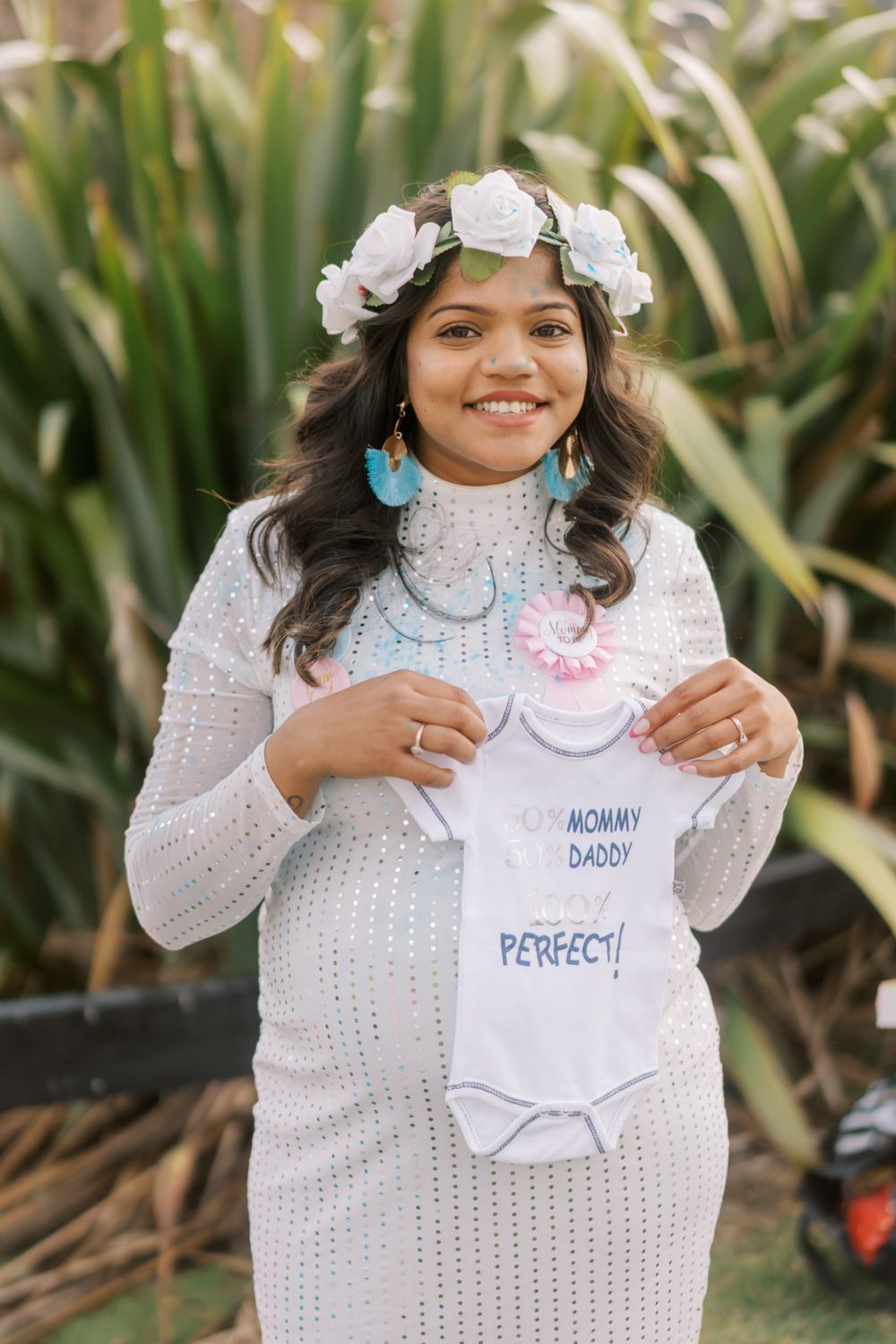 Smiling pregnant woman in white dress holding baby onesie with floral crown outdoors at baby shower.