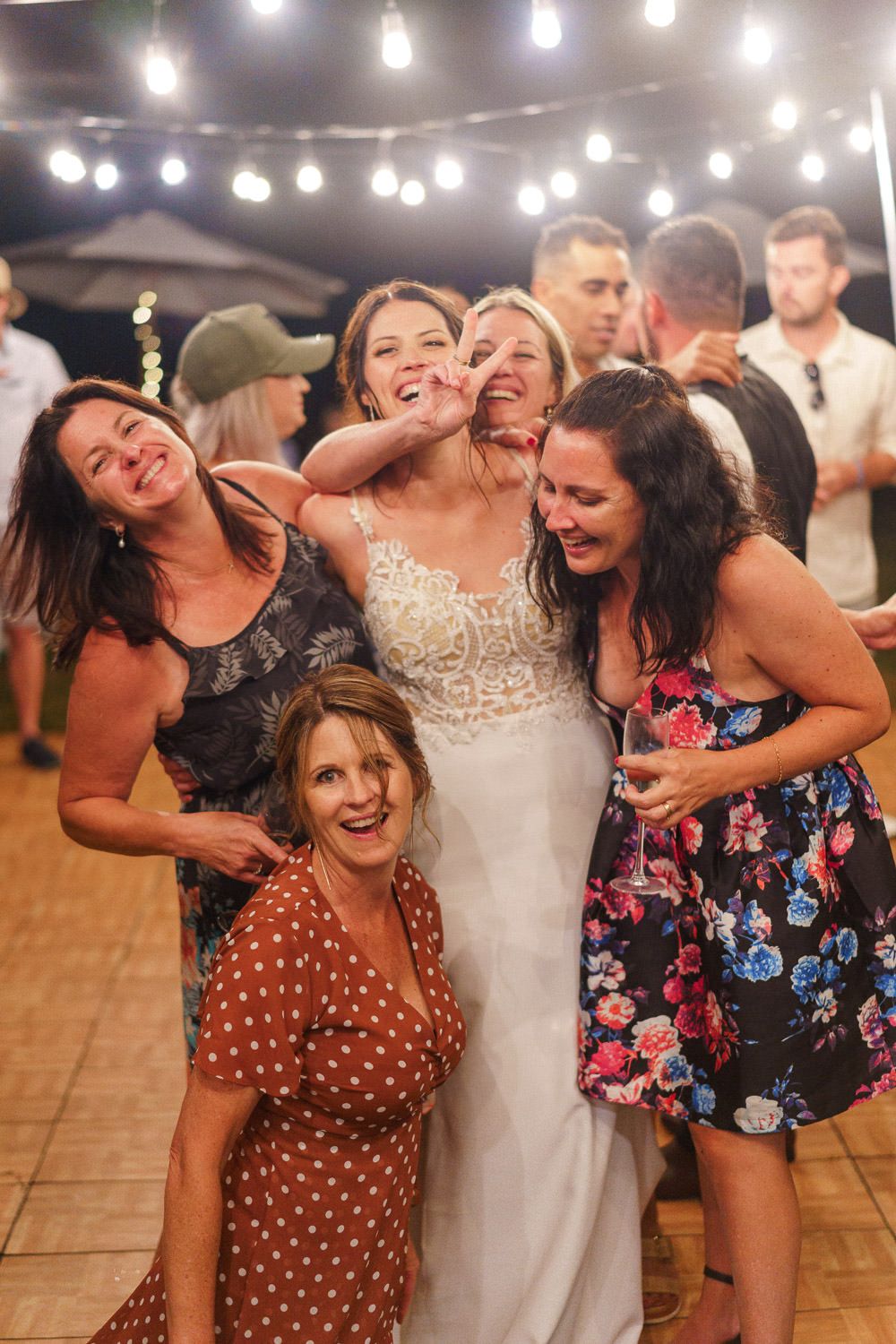 Bride in white lace gown laughs joyfully with female wedding guests under string lights at outdoor reception.
