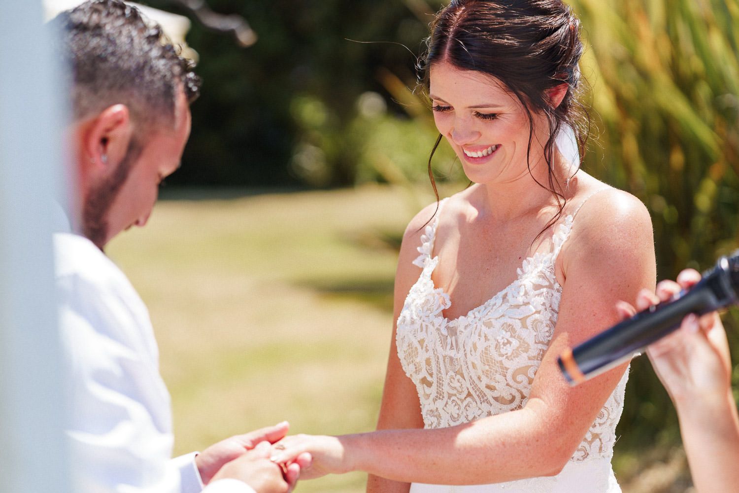 Bride smiling as groom places wedding ring on her finger during outdoor ceremony.