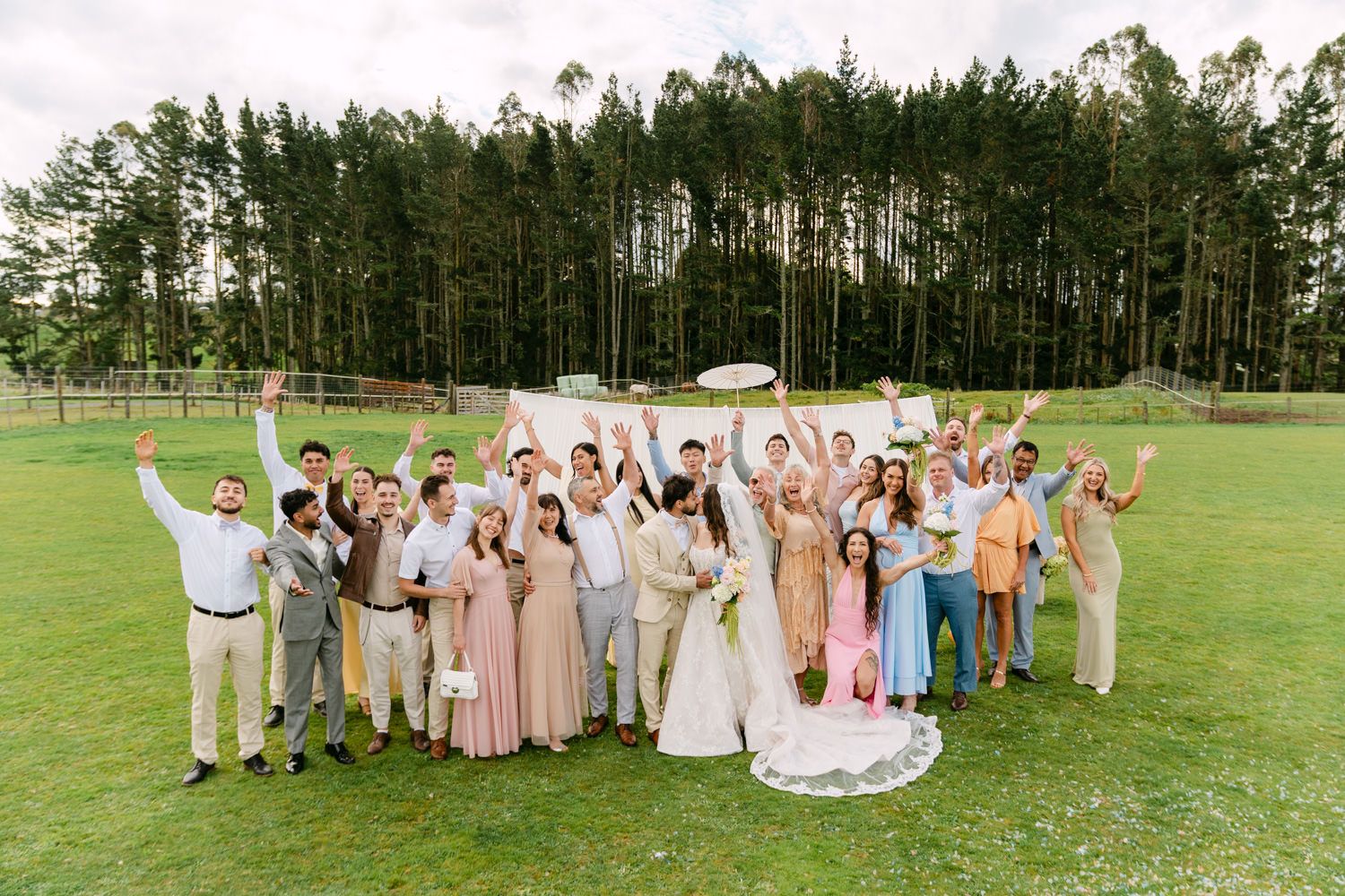 Large wedding party celebrating outdoors on green lawn with tall pine trees, bride in white gown surrounded by guests.