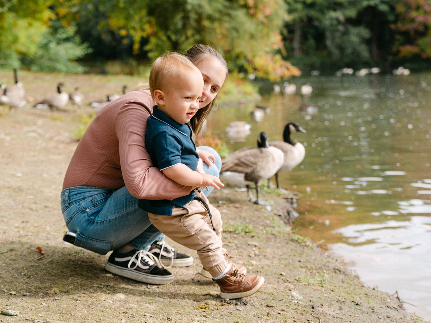 A mother crouches with her toddler by a pond, watching Canada geese near the water's edge in an autumn park.