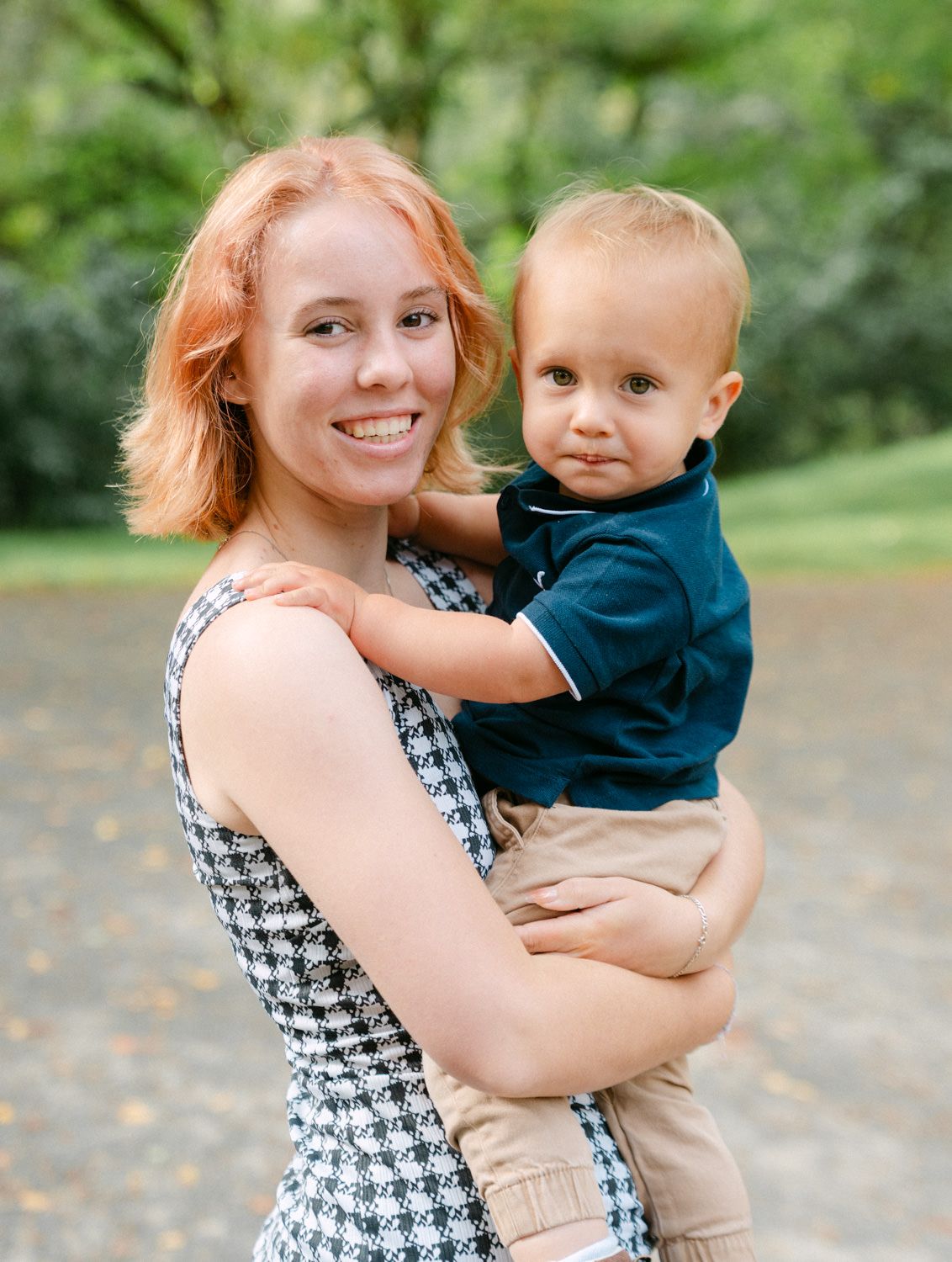 Smiling redhead woman holding a toddler boy in a navy polo outdoors in a lush green park setting.