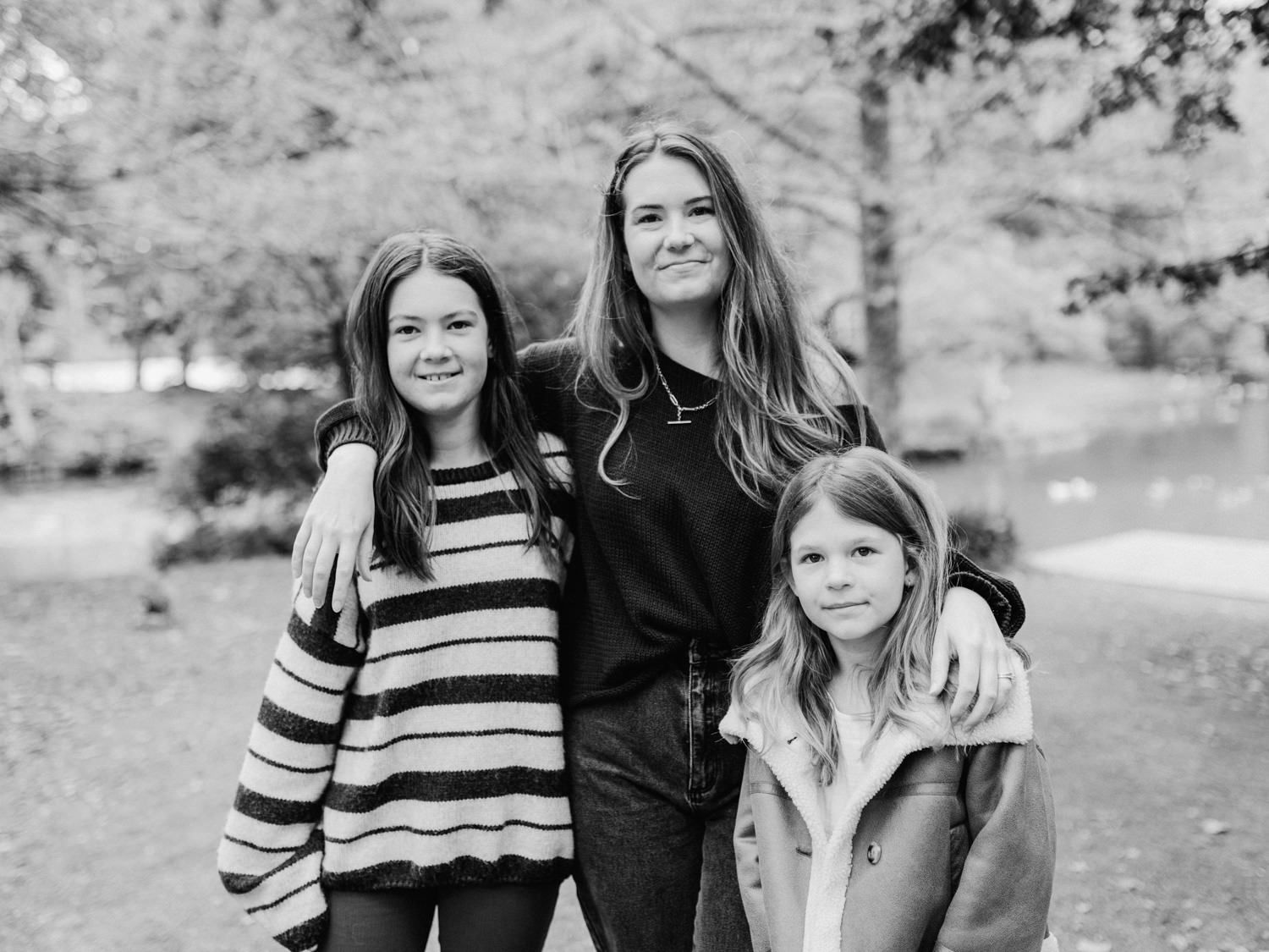Three girls pose together outdoors in a black and white photo, smiling warmly in a park setting.