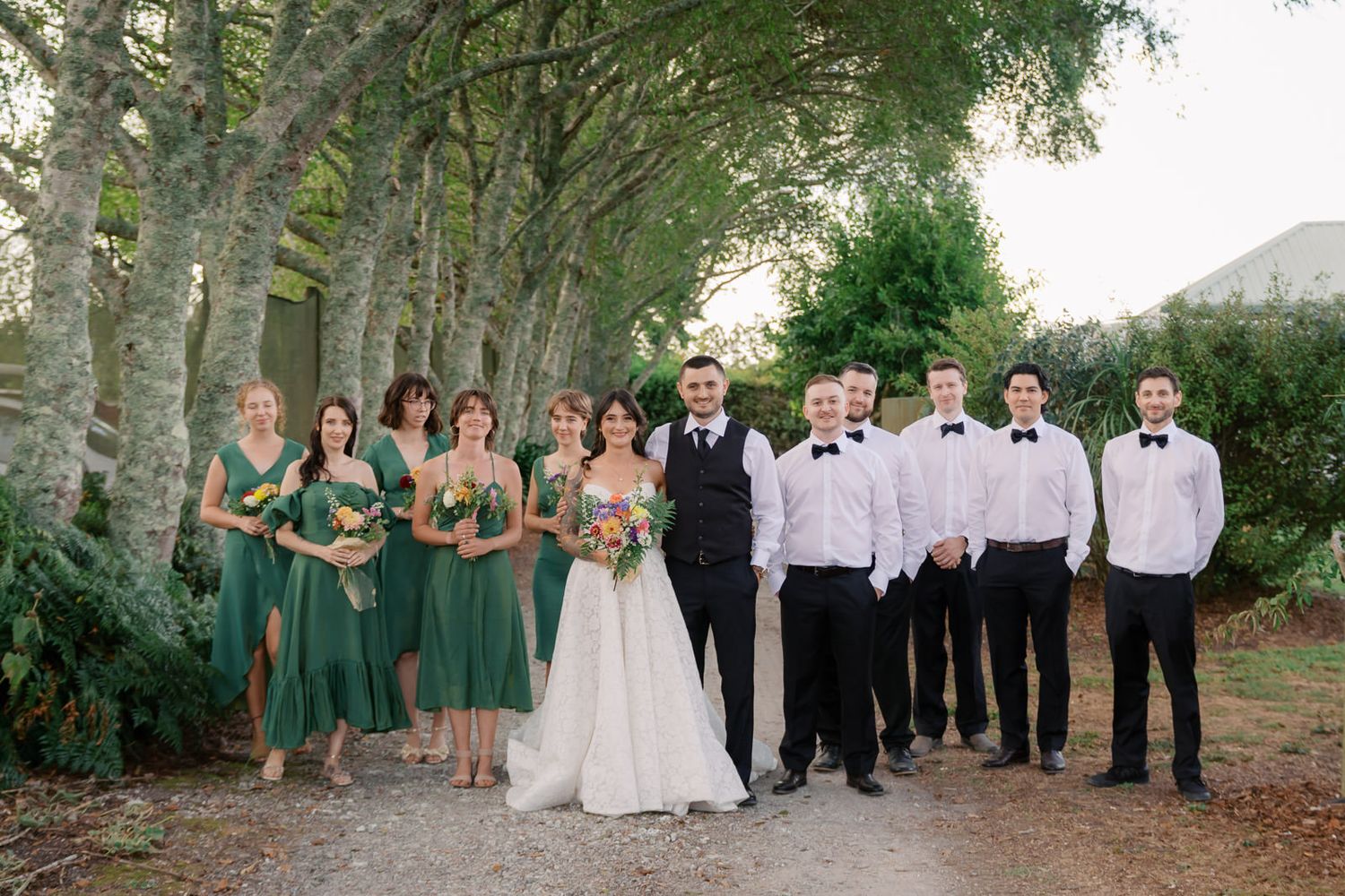 Wedding party posing outdoors with bridesmaids in green dresses and groomsmen in black bowties among tall trees.