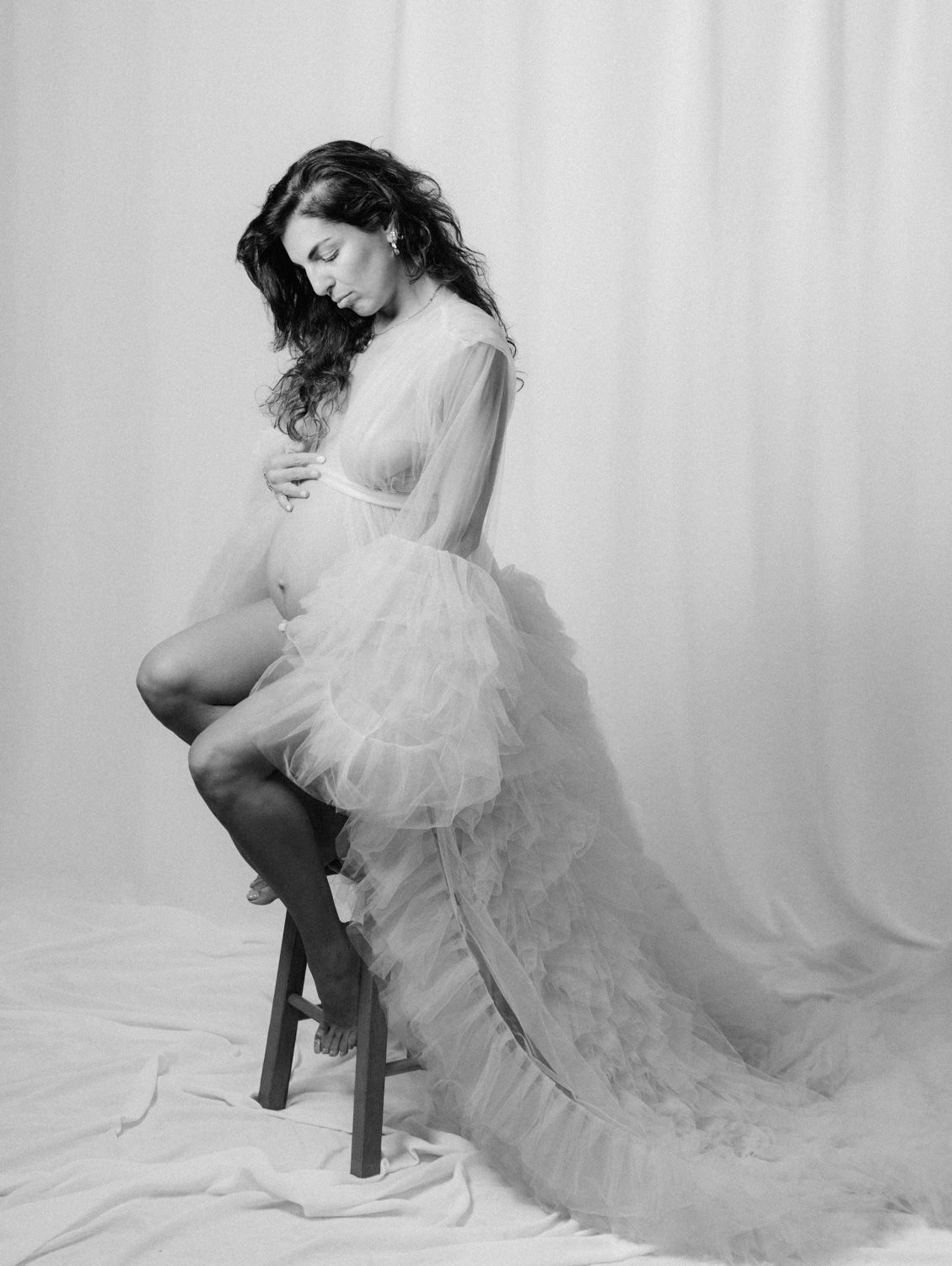 Black and white maternity photo of pregnant woman in sheer ruffled gown sitting gracefully on stool.