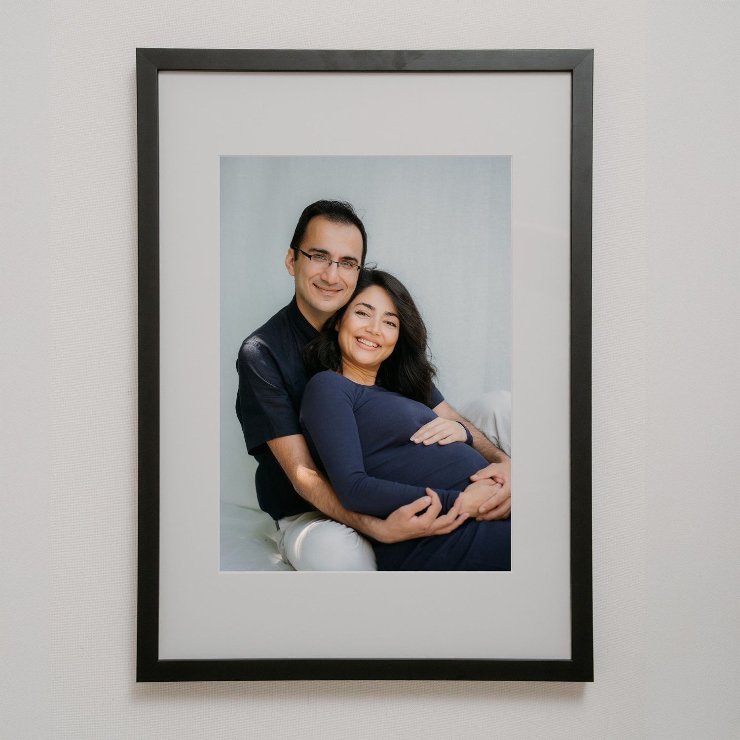 Framed maternity portrait of a smiling couple, man in black shirt embracing pregnant woman in navy dress, displayed on wall.