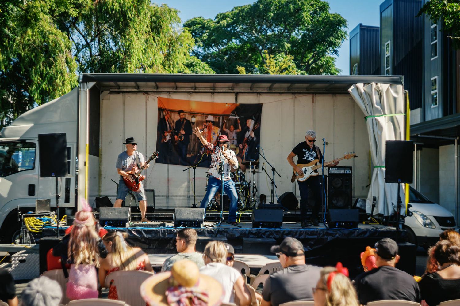 Live band performing on an outdoor mobile stage at a sunny festival with an audience gathered in front.