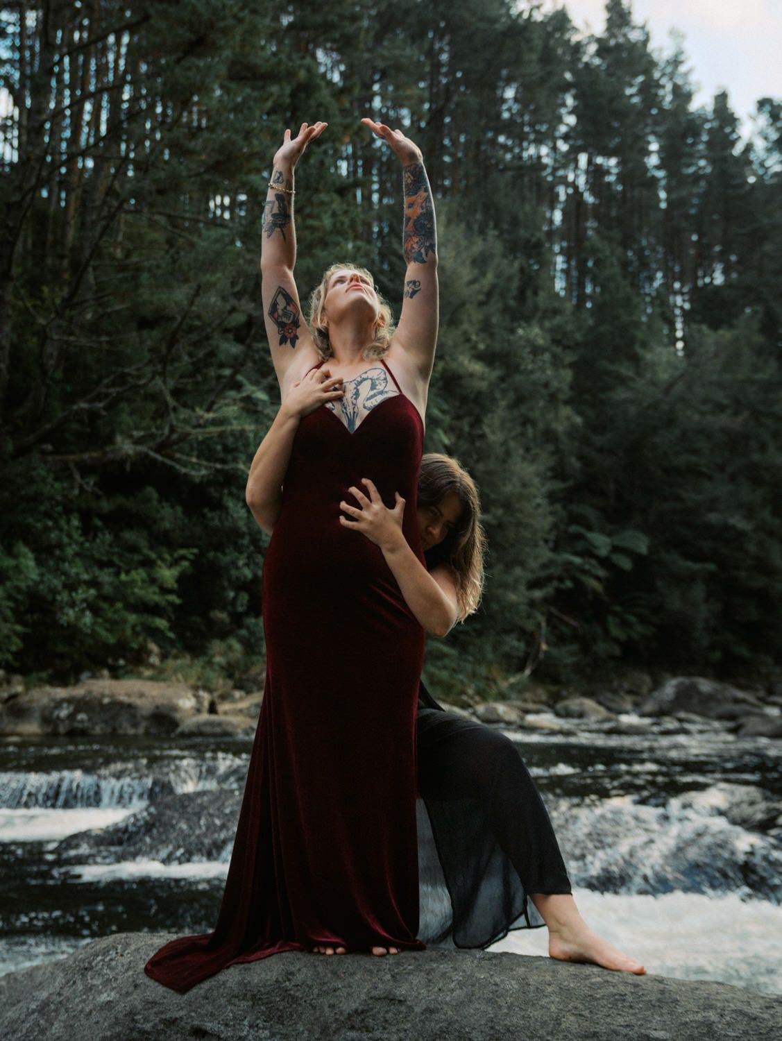 Two tattooed women pose dramatically on rocks by a waterfall, one in a burgundy gown with arms raised skyward.
