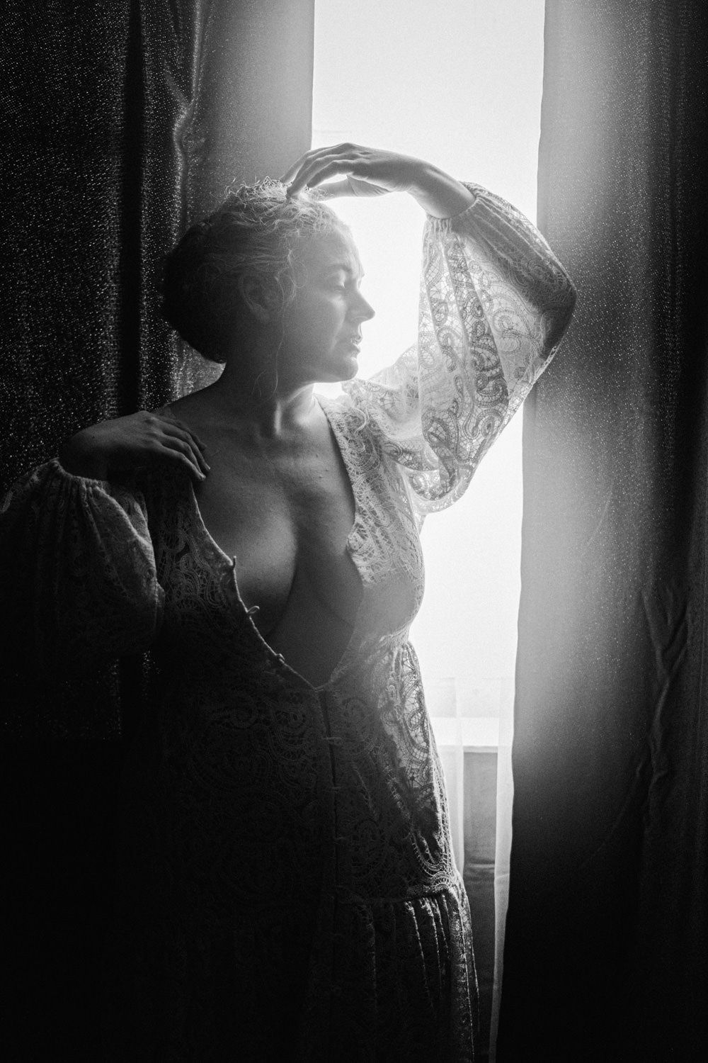 Black and white photo of a woman in lace dress standing by sunlit curtains, gazing thoughtfully out a bright window.