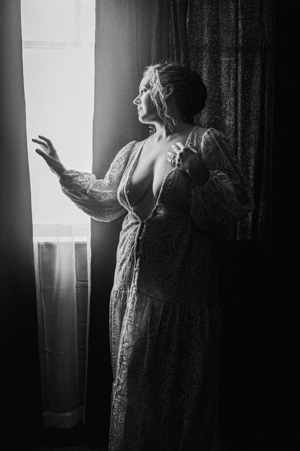 Black and white photo of a woman in a lace dress reaching toward a bright window with dramatic lighting.