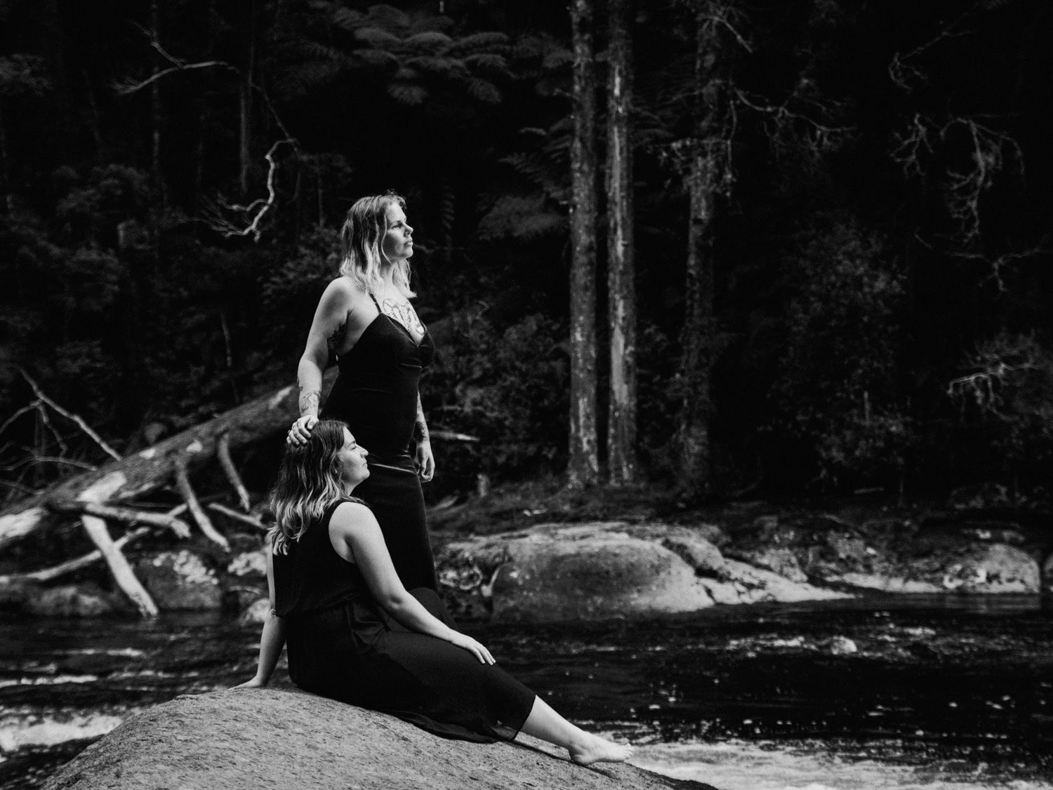 Two women in black attire pose gracefully on rocks by a forest stream in a serene black and white photo.