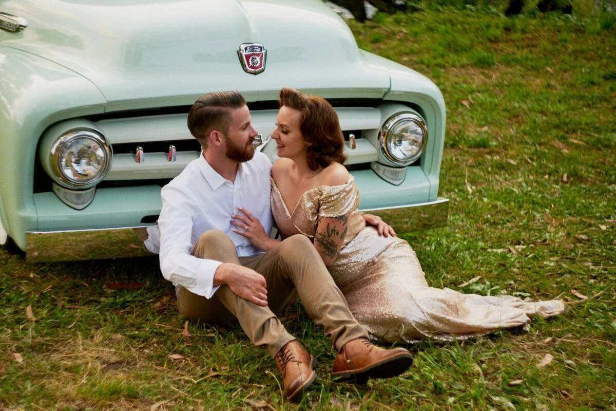 Couple sitting on grass leaning against a vintage mint green Ford truck during an outdoor engagement photo session.