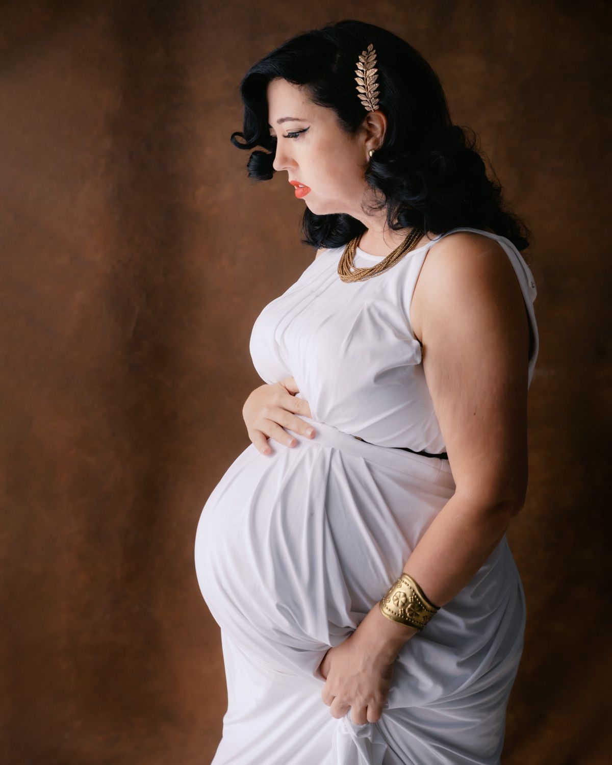 Pregnant woman in white Grecian dress with gold accessories poses gracefully in a studio maternity portrait.